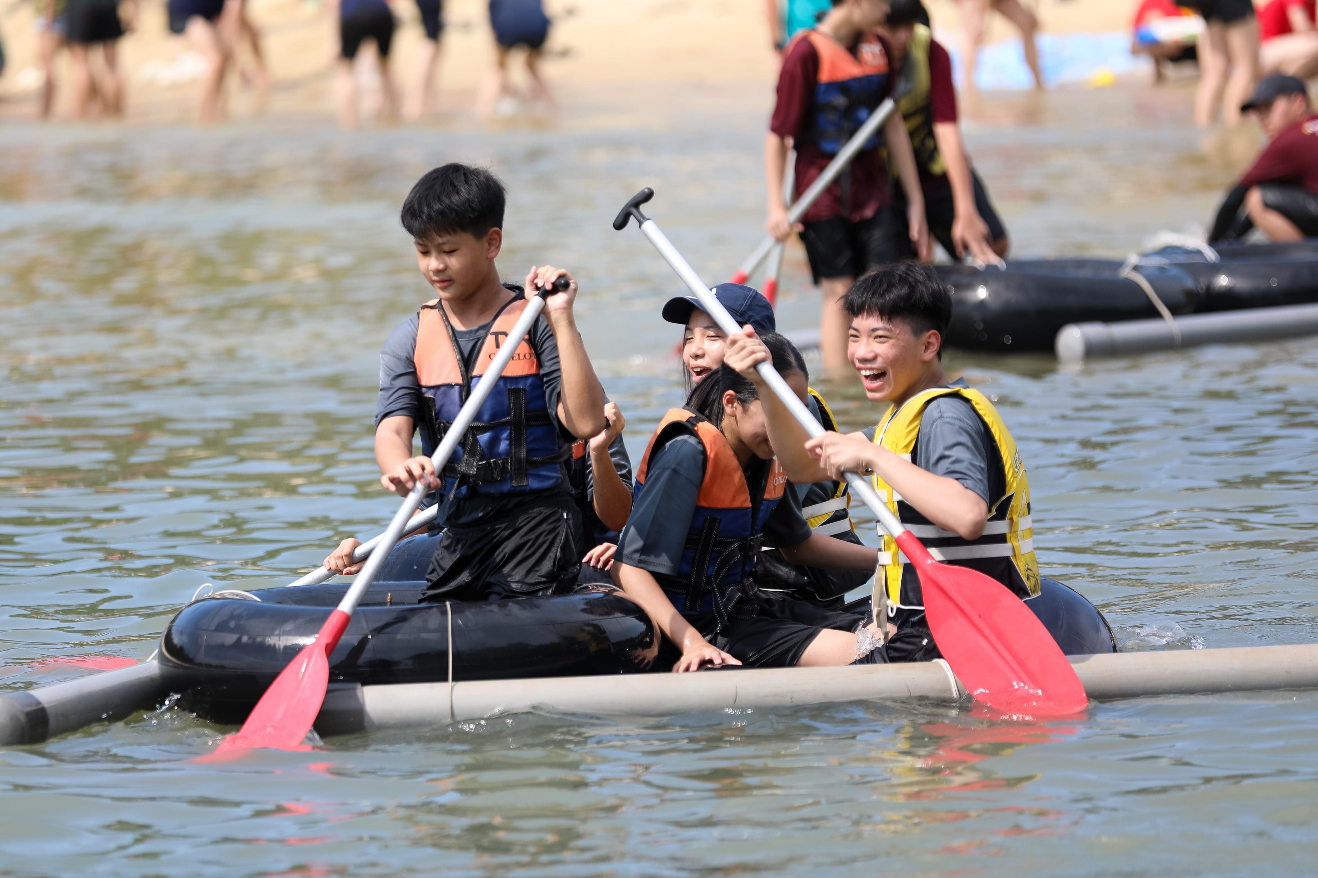 A group of students wearing life jackets smile and laugh as they work together to paddle a handmade raft constructed from inflatable inner tubes and plastic pipes in shallow water near a beach.